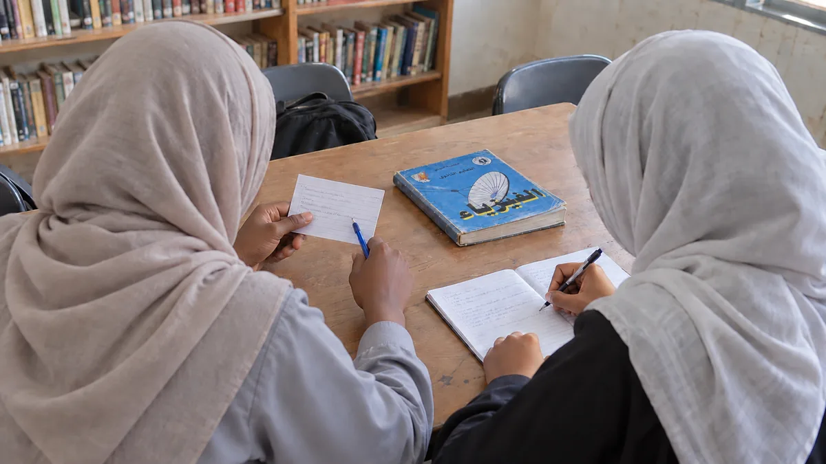 طالبتان من الخلف؛ إحداهما تسأل من بطاقة والأخرى تكتب الإجابة من ذاكرتها، والكتاب مغلق بينهما