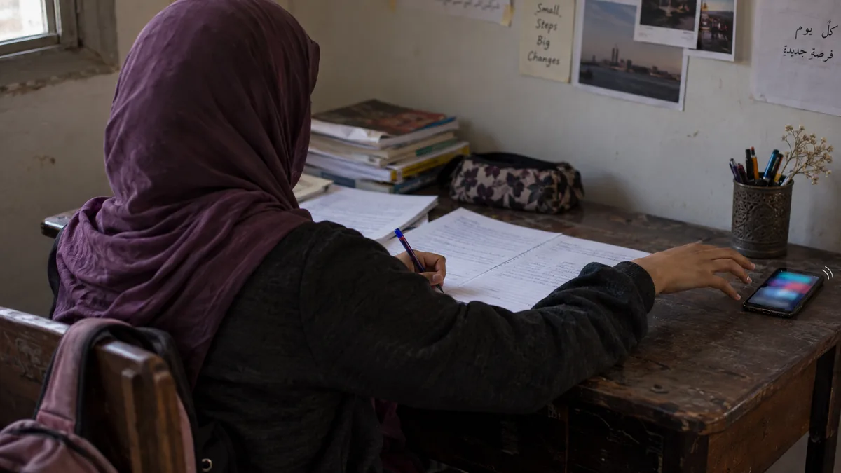 طالبة من الخلف تتوقف يدها عن الكتابة وتمتد نحو هاتف مضيء على طرف المكتب